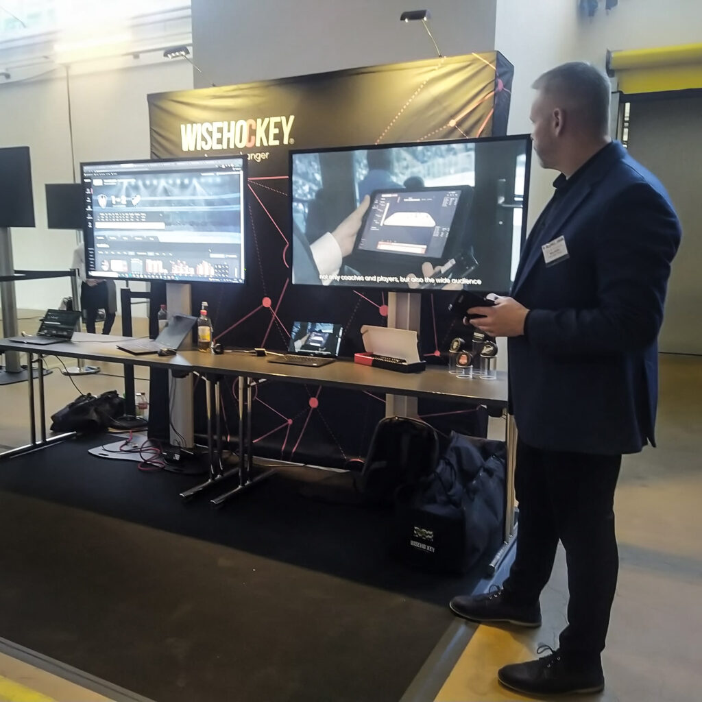 A stand with Wisehockey logo in the background and two monitors showing Wisehockey data. A man is standing next to the stand.