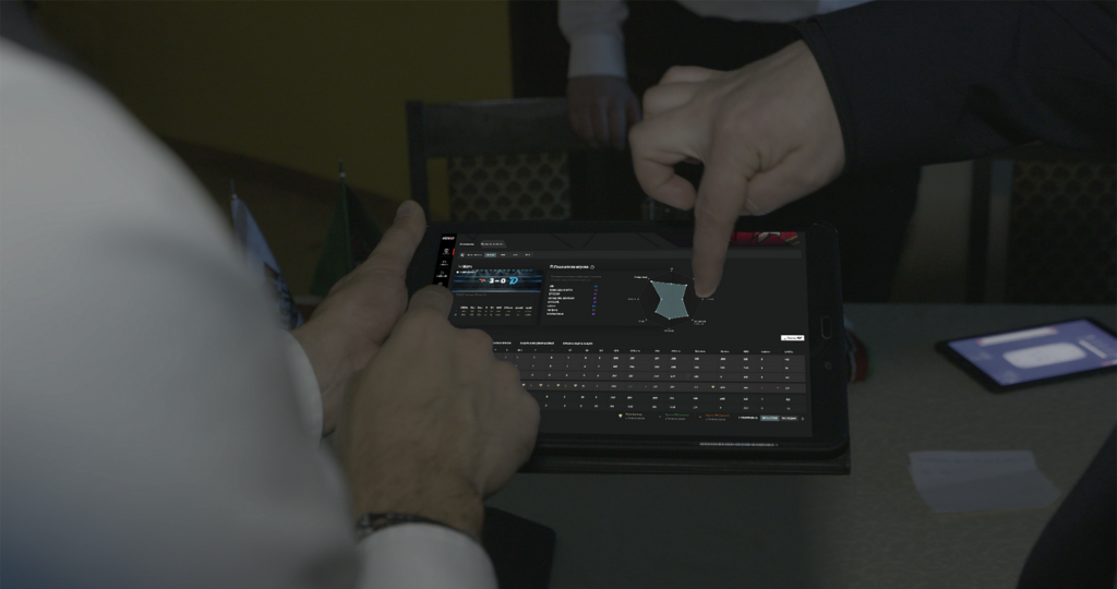 A member of team personnel is holding a tablet that shows a player's shift statistics. Two people are pointing at the statistics, presumably discussing them.