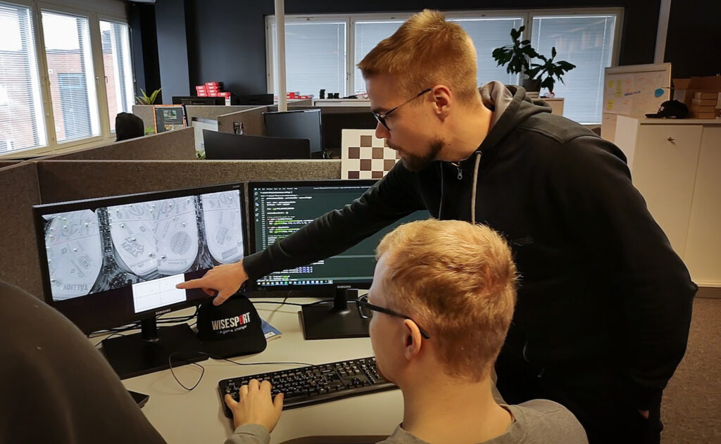Standing in the office, a man (Karjalainen) points at a computer screen. Another man is sitting at the desk watching the screen.